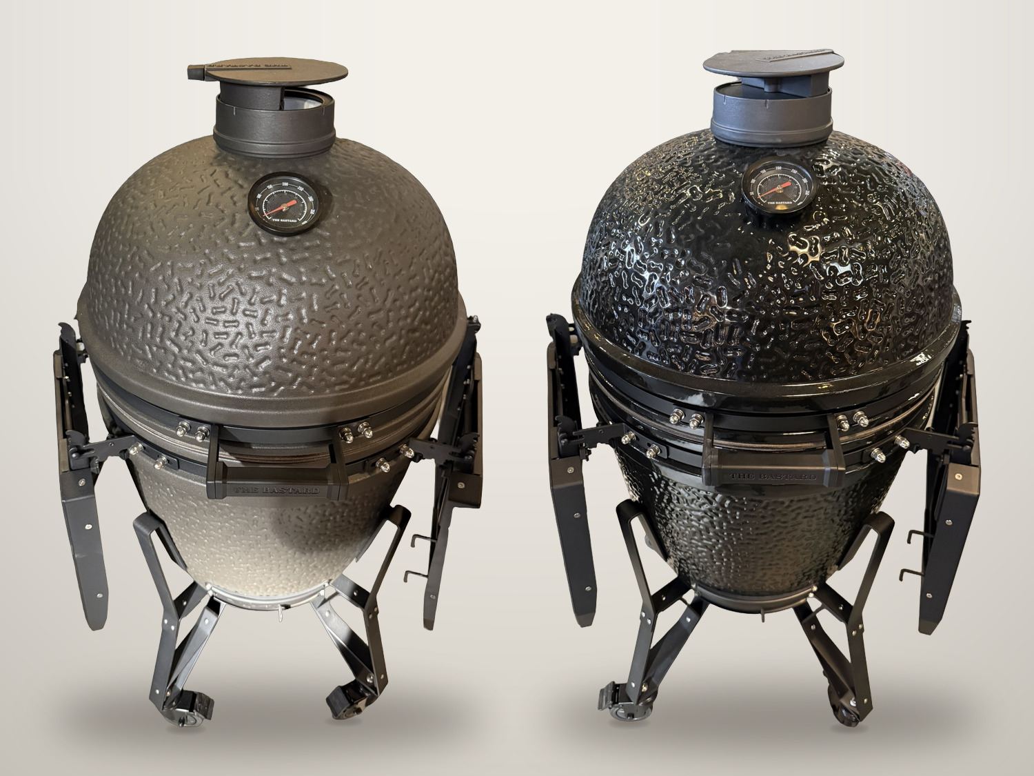 Twee The Bastard kamado's naast elkaar op een neutrale achtergrond: links de matte, ruwe uitvoering, rechts de hoogglans zwarte, allebei met gesloten deksel en ingeklapte zijtafels.